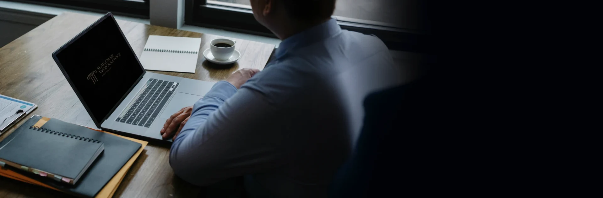 Adwokat podczas pracy przy biurku, na którym jest notatnik, laptop oraz kubek z kawą